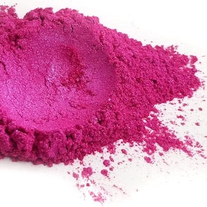 May include: A pile of vibrant, fuchsia-colored pigment powder against a white background. The powder has a metallic sheen, with a concentrated, smooth center and a textured, granular outer edge. The color is consistent throughout.