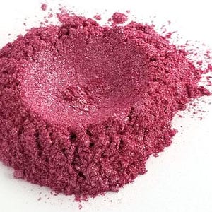 May include: A close-up of a pile of shimmering, deep rose-colored powder. The powder has a textured, granular appearance and reflects light, creating a sparkling effect. The powder is on a white background.