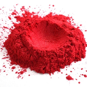 May include: A vivid red powder, possibly a pigment or dye, is scattered on a white surface. The powder is concentrated in a central mound with a crater-like depression, and smaller particles are spread around the edges.