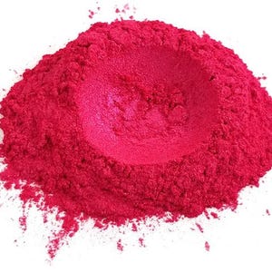 Puede incluir: Un montón de polvo de pigmento rosa intenso sobre un fondo blanco. El polvo tiene una apariencia texturizada con una depresión central y partículas dispersas en los bordes. El color es un rosa brillante y saturado.