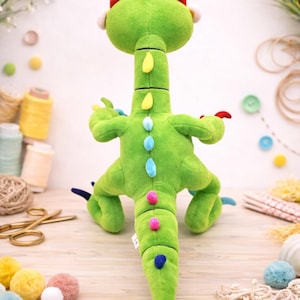 May include: A green plush dinosaur toy with a long neck and tail, featuring red, yellow, blue, and pink accents. The stuffed animal has a soft, textured appearance and is designed to stand upright. The background includes craft supplies and flowers.