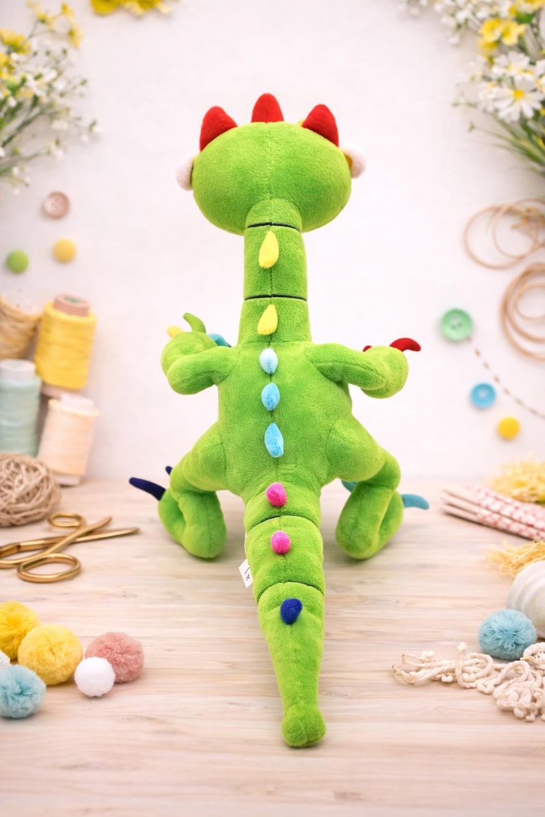 May include: A green plush dinosaur toy with a long neck and tail, featuring red, yellow, blue, and pink accents. The stuffed animal has a soft, textured appearance and is designed to stand upright. The background includes craft supplies and flowers.