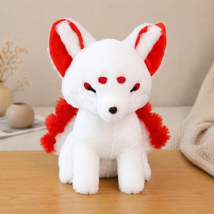 May include: A plush white fox with red accents on the ears and tail. The fox has red markings around the eyes and a brown nose. The stuffed animal is sitting on a wooden surface.