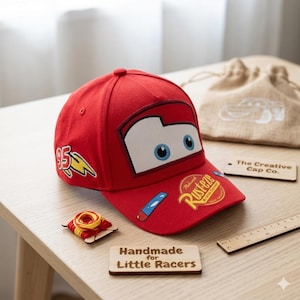May include: A red baseball cap featuring a cartoon car design with blue eyes and the number 95. The cap has a curved brim and the text "Rusteze" on the front. The cap is part of "The Creative Cap Co." collection. The text "Handmade for Little Racers" is also visible.