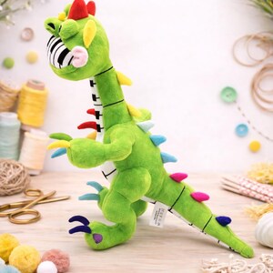 May include: A green plush dinosaur toy with a piano key-patterned mouth and colourful accents. The toy has red, yellow, blue, and pink details, and is designed to resemble a musical instrument. It is a whimsical and playful item.