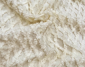 Puede incluir: Una tela de color crema con un patrón de diamantes texturizado y en relieve. La superficie está compuesta por hilos suaves y copetudos que forman un diseño geométrico de celosía repetitivo en todo el material, creando una apariencia táctil y dimensional.