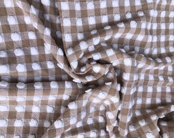 Puede incluir: Primer plano de una tela de cuadros vichy beige y blanca con un patrón de puntos texturizados y abultados. La tela está doblada y arrugada, mostrando la textura del material.