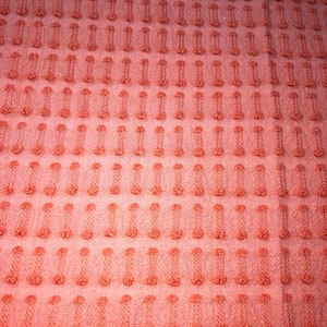 May include: A close-up of a coral-colored fabric with a textured pattern of small raised dots and vertical lines.