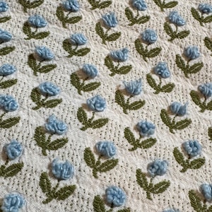 Könnte beinhalten: Nahaufnahme eines cremefarbenen Stoffes mit einem sich wiederholenden Muster aus blauen Pomponblumen und grün gestickten Stielen und Blättern. Der Stoff hat ein strukturiertes, gepunktetes Aussehen und erzeugt eine weiche, einladende Ästhetik.
