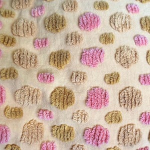 Può includere: Primo piano di un tessuto strutturato con un motivo ripetuto di forme trapuntate in rilievo. Le forme sono in tonalità di rosa e beige, sparse su uno sfondo color crema. Il design crea un effetto tattile e dimensionale.