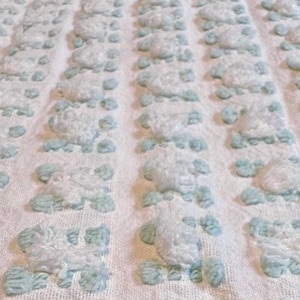 May include: Close-up of a white textured blanket with rows of raised, light blue floral-like designs. The blanket has a soft, plush appearance, suggesting comfort and warmth. The pattern is evenly spaced across the fabric.