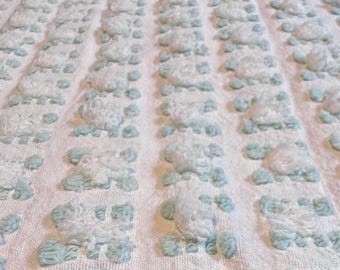 Puede incluir: Primer plano de una manta blanca texturizada con filas de diseños florales en relieve de color azul claro. La manta tiene un aspecto suave y afelpado, lo que sugiere comodidad y calidez. El patrón está espaciado uniformemente sobre la tela.