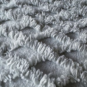 Könnte beinhalten: Nahaufnahme eines weißen, strukturierten Stoffes mit einem gewebten Muster. Der Stoff hat eine weiche, flauschige Textur und ist aus einem natürlichen Material gefertigt.