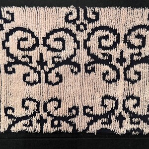 May include: A rectangular, textured bath mat with a cream-colored background and a repeating pattern of dark navy blue scrollwork. The mat has a soft, plush appearance and is likely made of absorbent material.