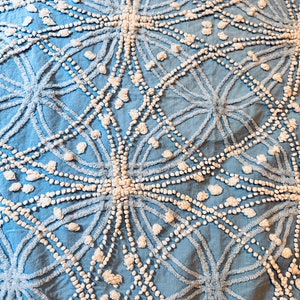 May include: A blue and white cotton throw blanket with a geometric pattern. The pattern is made up of circles and lines, and the circles are filled with small white pom-poms.