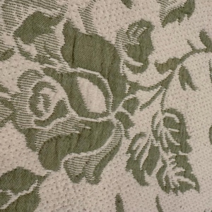 Pode incluir: Um tecido texturizado com um padrão floral repetitivo de rosas e folhas verde-sálvia tecidas num fundo branco sujo. O material apresenta uma trama distinta com uma textura de superfície em relevo em todo o design floral.