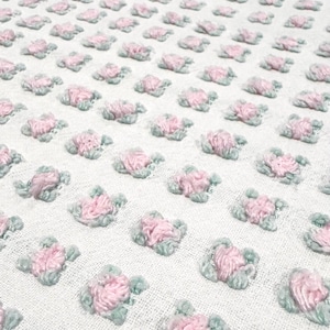以下が含まれることがあります： ピンクと淡い緑色の刺繍の花が繰り返される模様の白い生地のクローズアップ。花は密集して配置され、テクスチャのある表面を作り出しています。この花の模様は、このテキスタイルに繊細でフェミニンな印象を与えています。