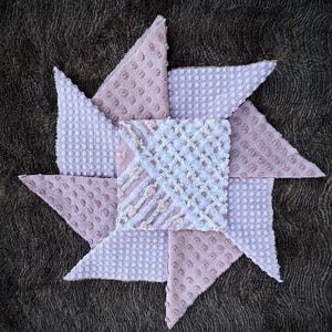 May include: A pink and white fabric pinwheel with a variety of textures and patterns. The pinwheel is made of eight triangular pieces of fabric, each with a different design. The pinwheel is arranged in a star shape.