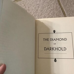 Puede incluir: Libro abierto con el t&iacute;tulo "THE DIAMOND OF DARKHOLD" en un recuadro negro sobre una p&aacute;gina color crema. El lomo del libro es amarillo y dorado. El libro est&aacute; abierto, revelando la p&aacute;gina del t&iacute;tulo.