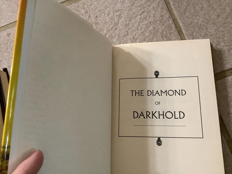Puede incluir: Libro abierto con el t&iacute;tulo "THE DIAMOND OF DARKHOLD" en un recuadro negro sobre una p&aacute;gina color crema. El lomo del libro es amarillo y dorado. El libro est&aacute; abierto, revelando la p&aacute;gina del t&iacute;tulo.