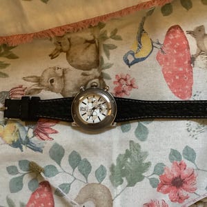 May include: A silver chronograph wristwatch with a black leather strap. The watch face has a white background with black markings and hands.