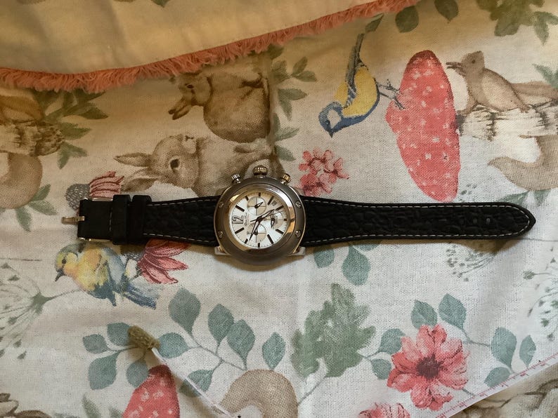 May include: A silver chronograph wristwatch with a black leather strap. The watch face has a white background with black markings and hands.