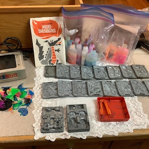 May include: A vintage toy set for making mini-dragons. The set includes a book titled "How to Make Mini-Dragons", a metal mold maker, plastic molds, colorful plastic pieces, and bottles of colored liquid.