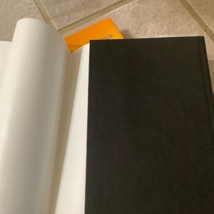 Puede incluir: Un libro abierto con p&aacute;ginas blancas y una cubierta negra. Las p&aacute;ginas est&aacute;n curvadas, revelando el interior. Una tarjeta naranja es visible en el fondo, descansando sobre una superficie embaldosada.