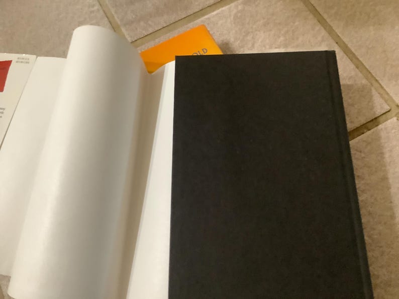 Puede incluir: Un libro abierto con p&aacute;ginas blancas y una cubierta negra. Las p&aacute;ginas est&aacute;n curvadas, revelando el interior. Una tarjeta naranja es visible en el fondo, descansando sobre una superficie embaldosada.