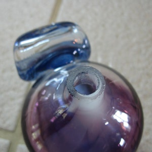 May include: A purple glass bottle with a blue glass stopper. The bottle has a round shape and a clear opening at the top.