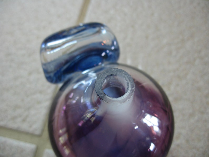 May include: A purple glass bottle with a blue glass stopper. The bottle has a round shape and a clear opening at the top.