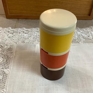 Puede incluir: Un recipiente apilado con tapa crema, sección media amarilla, centro naranja y base marrón. El recipiente está sobre un paño blanco con borde de encaje.