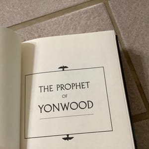 Puede incluir: Libro abierto con el t&iacute;tulo "THE PROPHET OF YONWOOD" en texto negro sobre una p&aacute;gina blanca. El t&iacute;tulo est&aacute; enmarcado por un rect&aacute;ngulo negro con iconos decorativos de p&aacute;jaros. El libro est&aacute; sobre una superficie clara.
