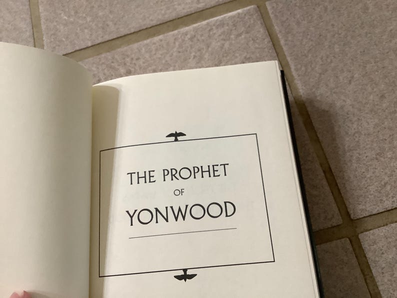 Puede incluir: Libro abierto con el t&iacute;tulo "THE PROPHET OF YONWOOD" en texto negro sobre una p&aacute;gina blanca. El t&iacute;tulo est&aacute; enmarcado por un rect&aacute;ngulo negro con iconos decorativos de p&aacute;jaros. El libro est&aacute; sobre una superficie clara.