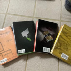 Puede incluir: Cuatro libros con portadas &uacute;nicas est&aacute;n dispuestos sobre una superficie embaldosada. Las portadas muestran varios dise&ntilde;os, incluyendo patrones abstractos, formas geom&eacute;tricas y una imagen celestial. Cada libro tiene una etiqueta de c&oacute;digo de barras.
