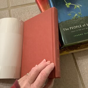 Puede incluir: Una pila de libros, incluyendo un libro rojo de tapa dura titulado "The City of Ember" y un libro azul y verde titulado "The People of Sparks". Los libros est&aacute;n sobre una superficie embaldosada. Una mano sostiene el libro rojo.
