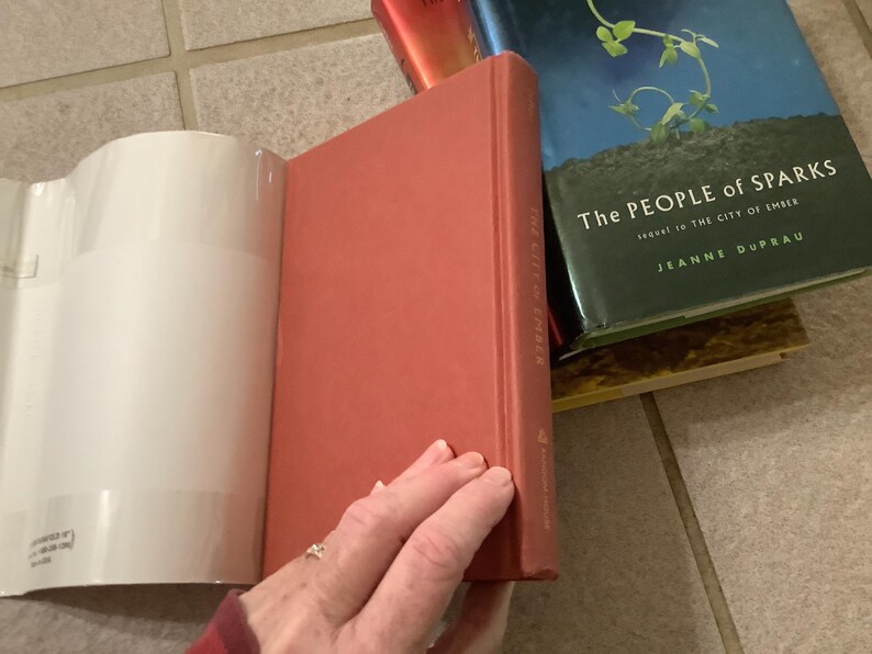 Puede incluir: Una pila de libros, incluyendo un libro rojo de tapa dura titulado "The City of Ember" y un libro azul y verde titulado "The People of Sparks". Los libros est&aacute;n sobre una superficie embaldosada. Una mano sostiene el libro rojo.