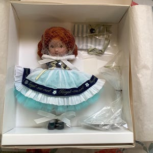 Puede incluir: Una muñeca de porcelana con pelo rojo que lleva un vestido azul y blanco con el texto "Forget-me-not" en la cinta. La muñeca está en una caja blanca con papel de seda.