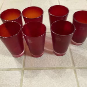 May include: Set of seven red-tinted glass tumblers. The glasses have a cylindrical shape with a slightly flared top. The interior of the glasses has an orange hue. The glasses are arranged on a tiled surface.