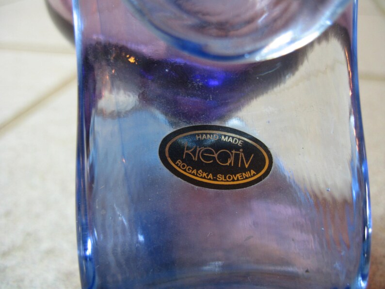 May include: A blue glass vase with a label that reads "Hand Made Kreativ Rogaska-Slovenia".