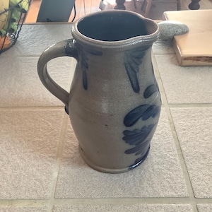 Puede incluir: Una jarra de gres con asa y boca ancha. La jarra es de color gris claro con diseños florales azules. La jarra está sobre una superficie de baldosas gris claro.