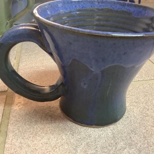Peut inclure: Mug en céramique fait à la main avec une forme unique et une grande anse. La tasse présente un glaçage bicolore, avec un bleu profond sur la partie supérieure et un bleu-vert plus foncé sur la partie inférieure. La tasse mesure environ 10 cm de haut.