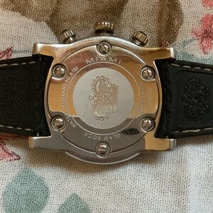 May include: A silver-toned watch with a black leather band. The watch face is white with a crown logo and the words "Glam Rock Miami" printed on it. The back of the watch has the words "Water Resistant 10 ATM" printed on it.