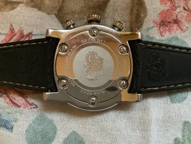 May include: A silver-toned watch with a black leather band. The watch face is white with a crown logo and the words "Glam Rock Miami" printed on it. The back of the watch has the words "Water Resistant 10 ATM" printed on it.