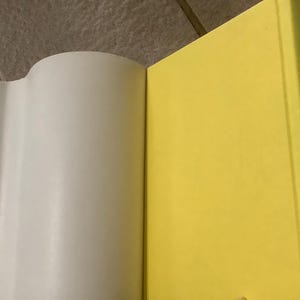 Puede incluir: Un libro abierto con p&aacute;ginas blancas y una cubierta amarilla brillante. El lomo del libro es visible, con el t&iacute;tulo "The Diamond of Darkhold" en texto azul. El libro est&aacute; sobre una superficie embaldosada.