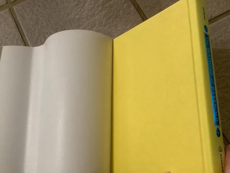 Puede incluir: Un libro abierto con p&aacute;ginas blancas y una cubierta amarilla brillante. El lomo del libro es visible, con el t&iacute;tulo "The Diamond of Darkhold" en texto azul. El libro est&aacute; sobre una superficie embaldosada.