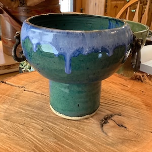 Puede incluir: Un macetero de cerámica verde con un esmalte azul y dos asas. El macetero está sobre un soporte de madera.