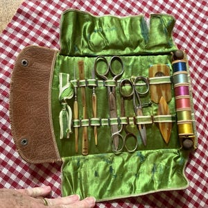 May include: An open antique sewing kit with a brown leather flap and a green velvet interior. The kit contains scissors, needles, spools of thread, and other sewing tools. The spools of thread are in various colors.