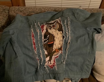 Könnte beinhalten: Eine blaue Jeansjacke mit einem großen gestickten Eulenmotiv auf der Rückseite. Die Jacke wurde mit Rissen und Löchern bearbeitet, was ihr einen Vintage-Look verleiht.