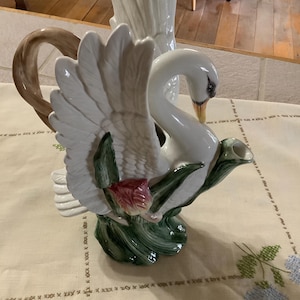 May include: A white ceramic swan-shaped teapot with a brown handle and a long spout. The swan has outstretched wings and is decorated with green leaves and a red flower. The teapot is on a cream-colored tablecloth.